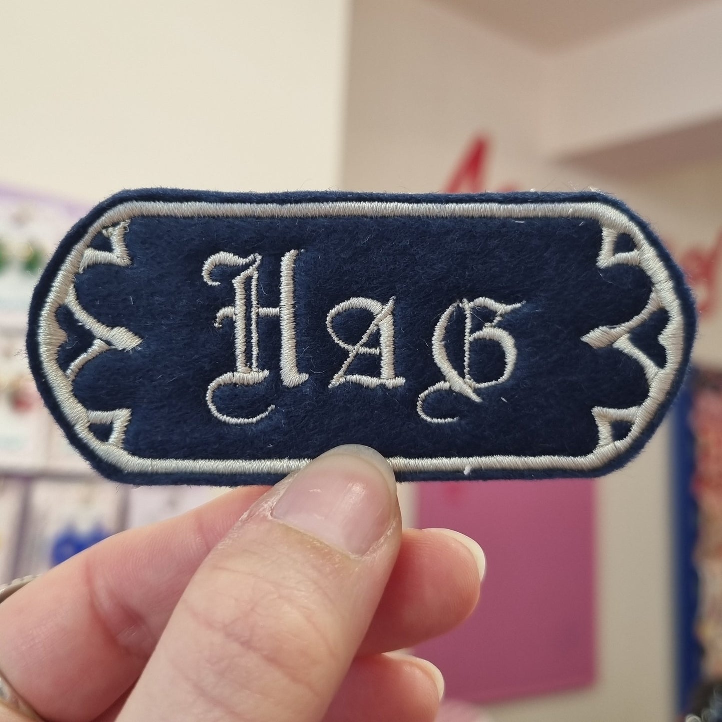 A navy sew-on patch held up between thumb and forefinger, has HAG written in medieval font