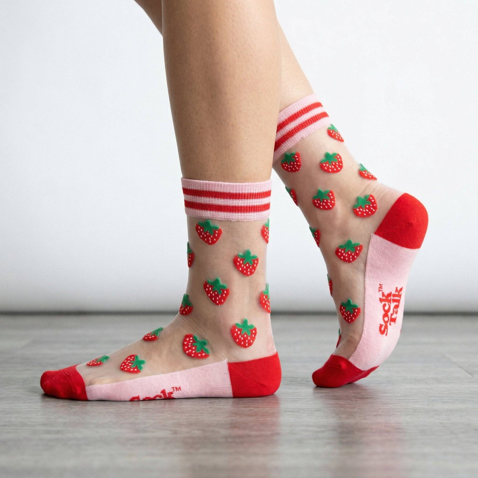 Sheer socks with strawberry pattern and red bottoms worn by a person on a light wooden floor.