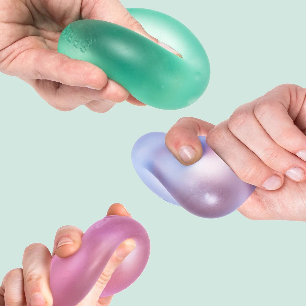 Three hands holding different translucent silicone stress balls in mint green, lavender, and pink against a pale mint background.