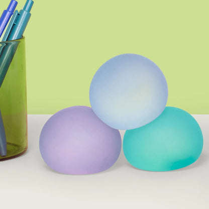Three pastel-colored silicone stress balls stacked together on a white surface, with a green pencil cup containing pens on the left, set against a light green background.