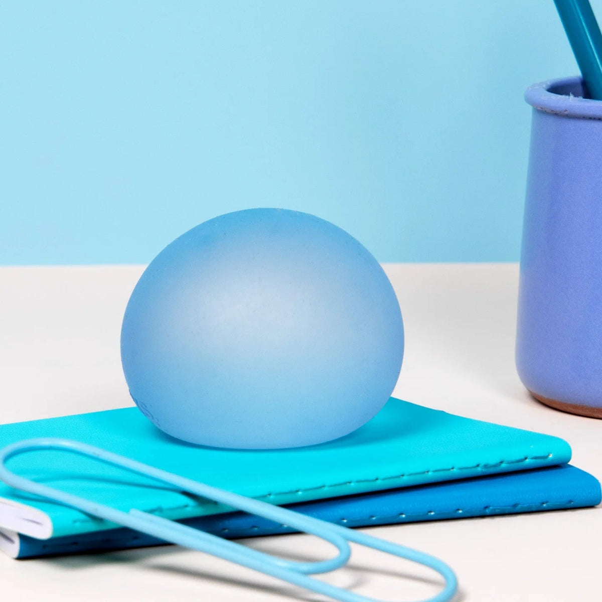 A light blue frosted glass sphere resting on a stack of teal and blue notebooks, with a matching light blue paperclip in the foreground and a blue pen holder on a white surface against a light blue background.