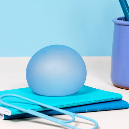 A light blue frosted glass sphere resting on a stack of teal and blue notebooks, with a matching light blue paperclip in the foreground and a blue pen holder on a white surface against a light blue background.