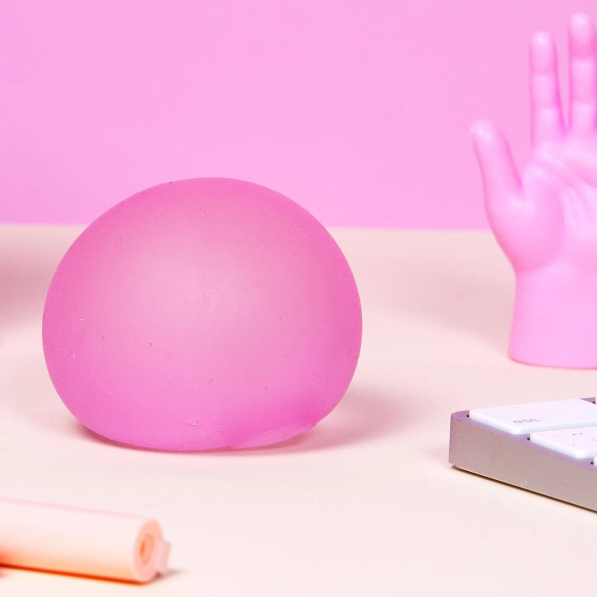 A matte pink spherical object on a light beige surface with a pink background, alongside a pink hand-shaped object, a small rectangular box, and a rolled pink item.