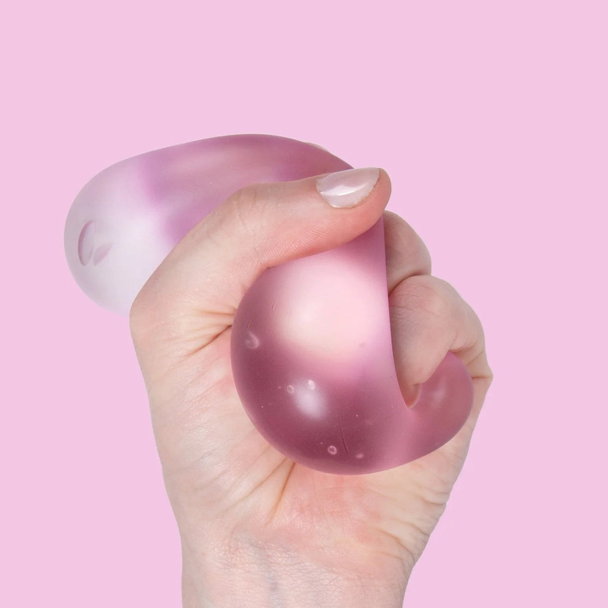 A hand squeezing a translucent pink stress ball against a solid pink background.