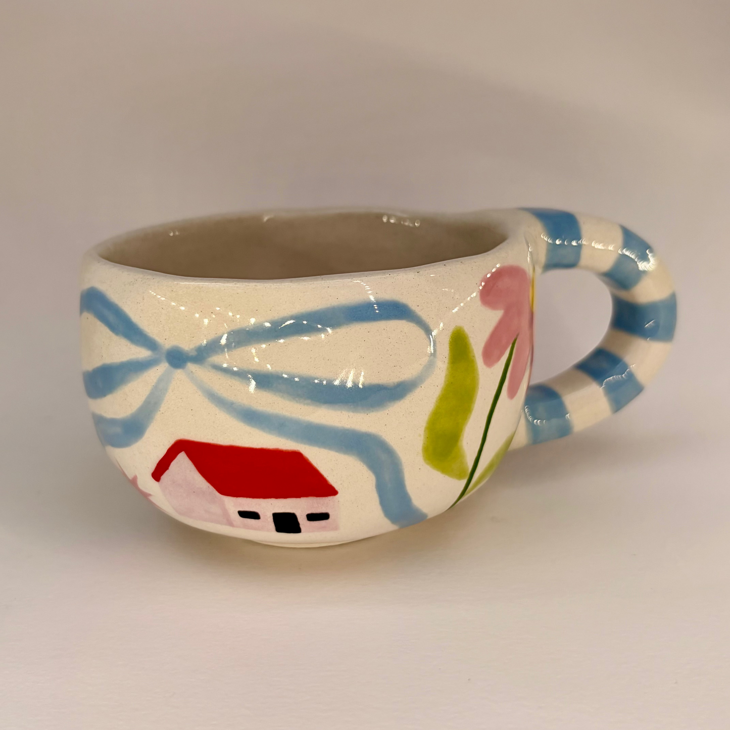 Ceramic mug with decorative handle featuring a house and flowers on a plain background