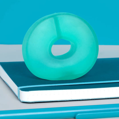 A translucent aqua-green donut-shaped silicone object resting on a blue notebook, with a matching blue pen in the foreground against a bright blue and gray background.
