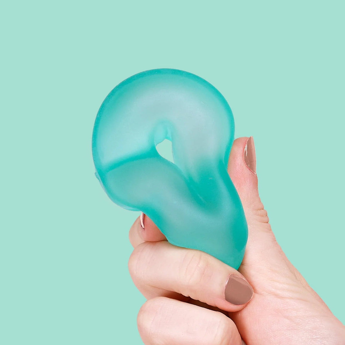 A hand with neutral nail polish holds a translucent aqua-green silicone stress toy against a solid mint green background.