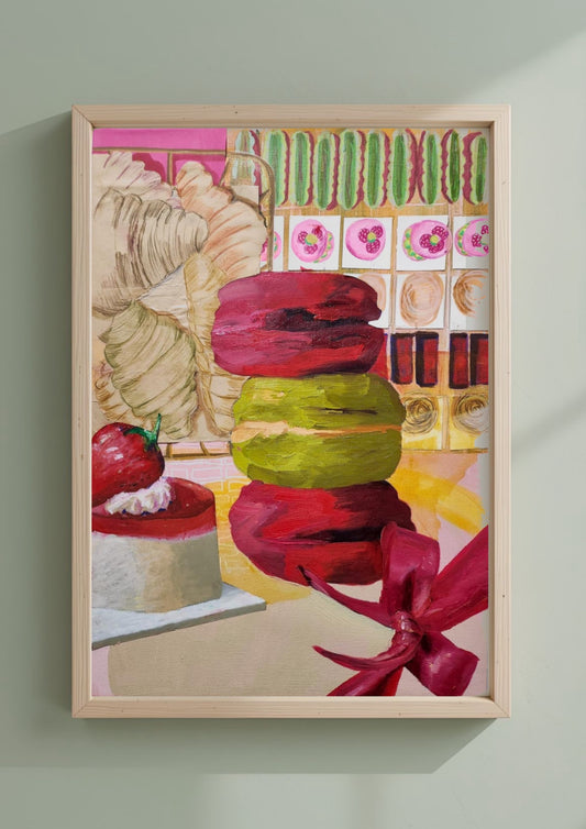A framed print of stacked macarons, a strawberry cake, and sweets, hanging on a light sage green wall.