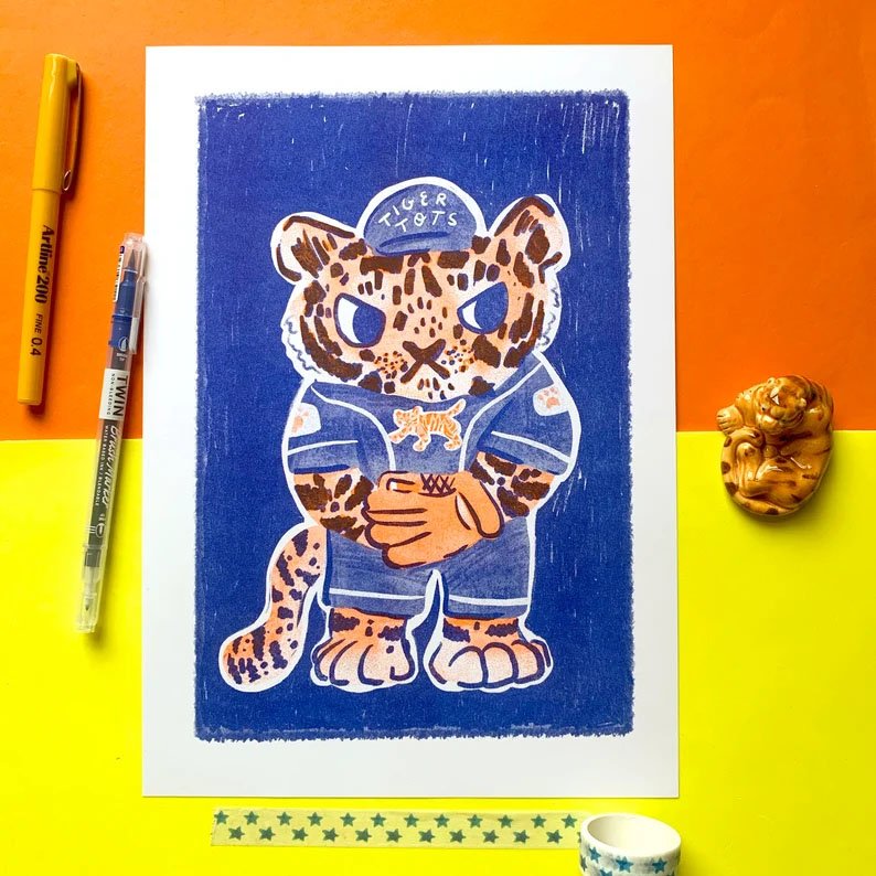 An art print featuring a cartoon tiger mascot in a blue baseball uniform, photographed from above on a bright yellow and orange surface with pens, washi tape, and a small figurine around it.