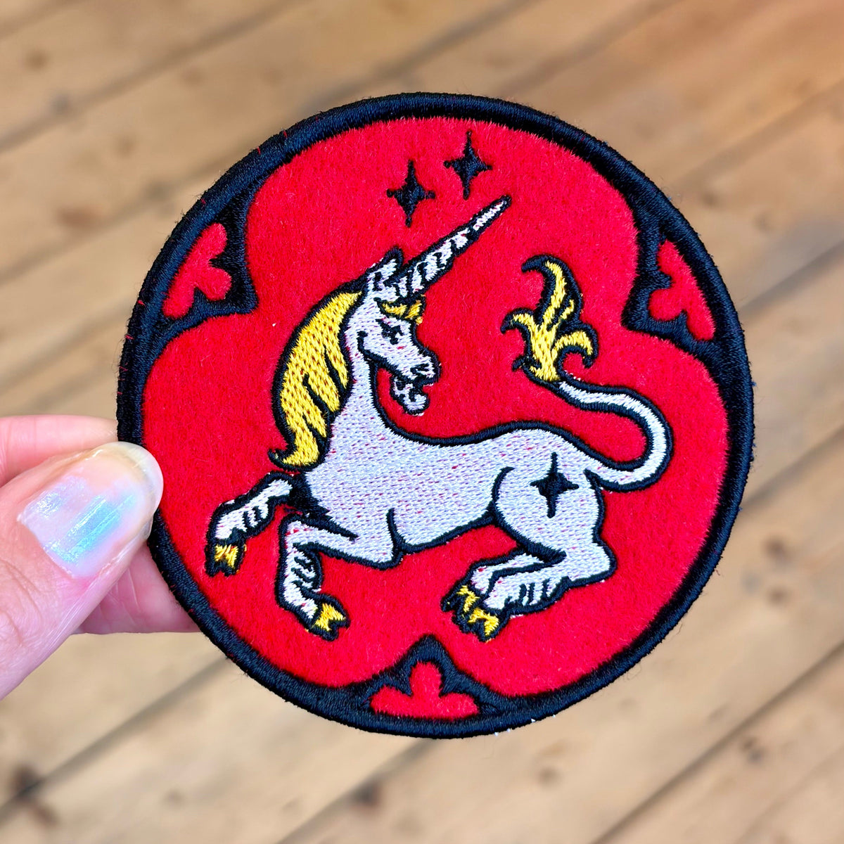 A circular embroidered patch featuring a white unicorn with a yellow horn and mane on a bright red background, held by a hand against a blurred wooden floor backdrop.