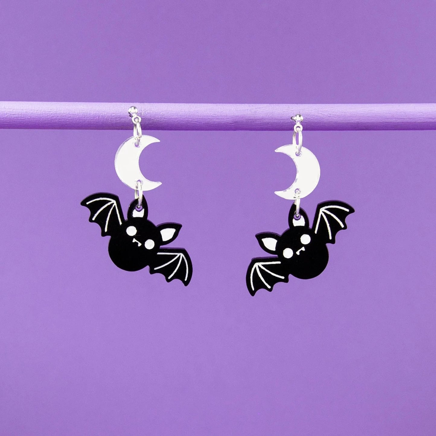 Bat-shaped earrings hanging from white moon pendants against a purple background