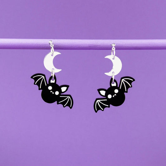 Bat-shaped earrings hanging from white moon pendants against a purple background