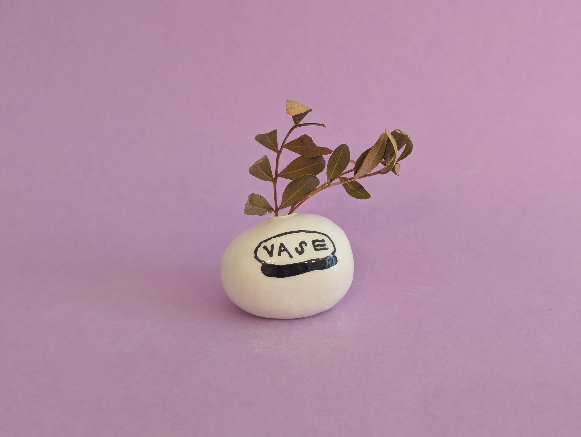 A small white ceramic vase with the word 'VASE' painted on it, holding a leafy sprig, set against a solid lavender background.