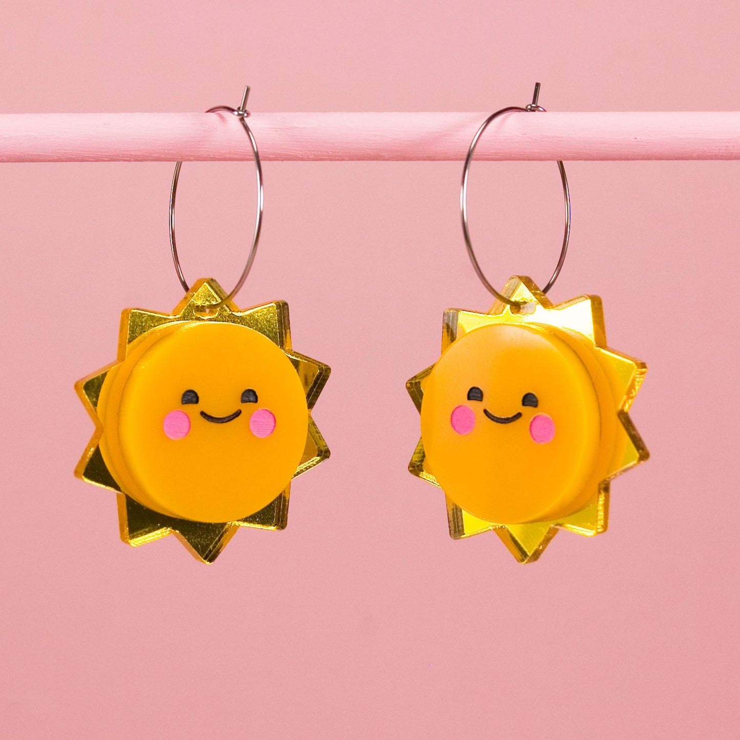Sun-shaped earrings with acrylic smiling faces, hanging from stainless steel hoops against a pink backgroundf