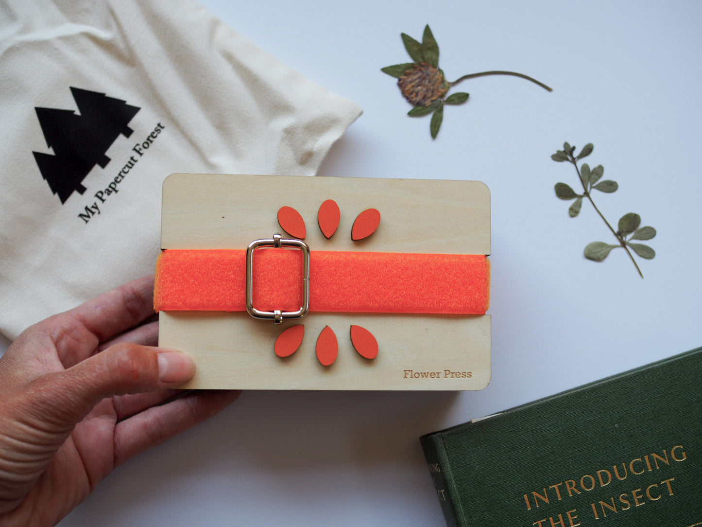 A hand holds a wooden flower-press with a bright orange velcro buckle across the centre. Against a background featuring pressed flowers, a tote carry bag, and a green book.