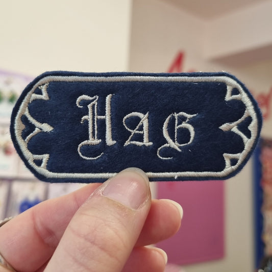 A navy sew-on patch held up between thumb and forefinger, has HAG written in medieval font
