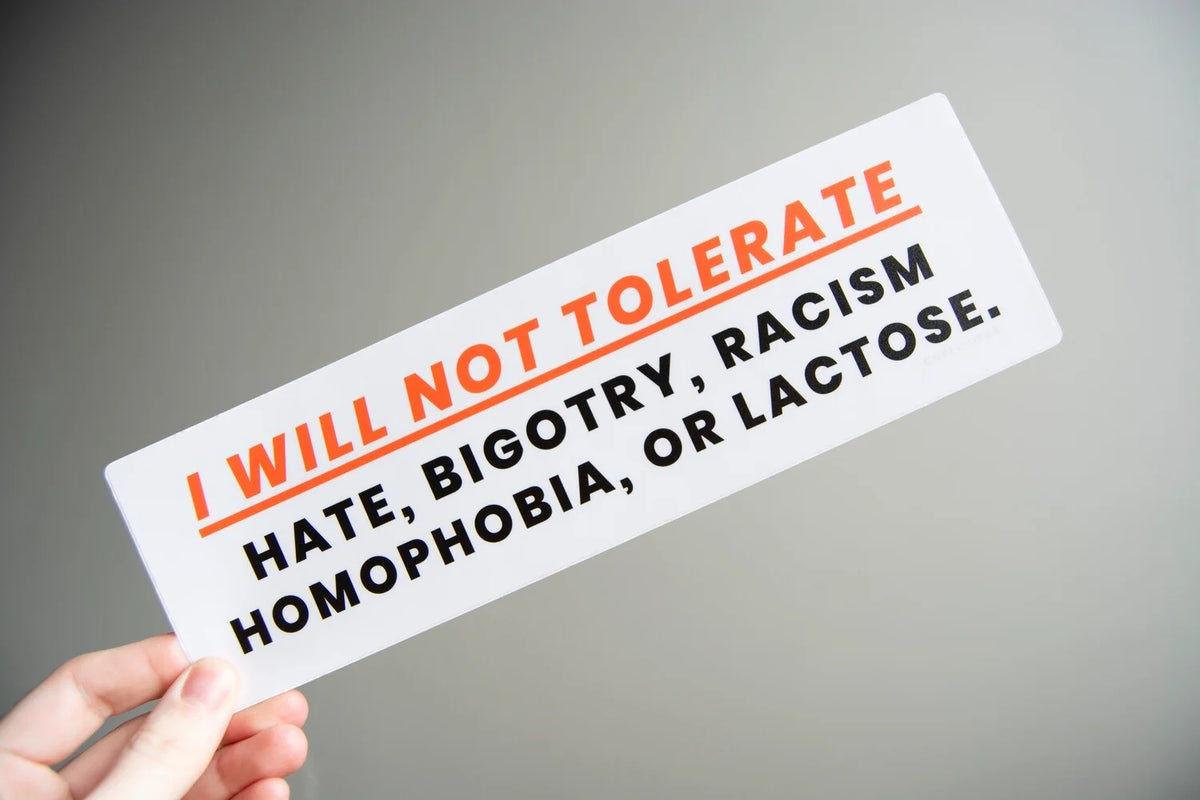 A hand holds a white rectangular bumper sticker with the text 'I WILL NOT TOLERATE HATE, BIGOTRY, RACISM HOMOPHOBIA, OR LACTOSE.' in orange and black lettering against a neutral gray background.