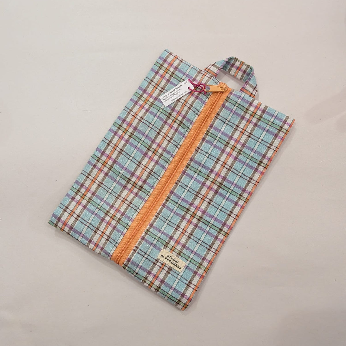 A folded plaid garment bag with a pastel multicolor pattern and a prominent orange zipper, shown on a plain light background with its tags attached.
