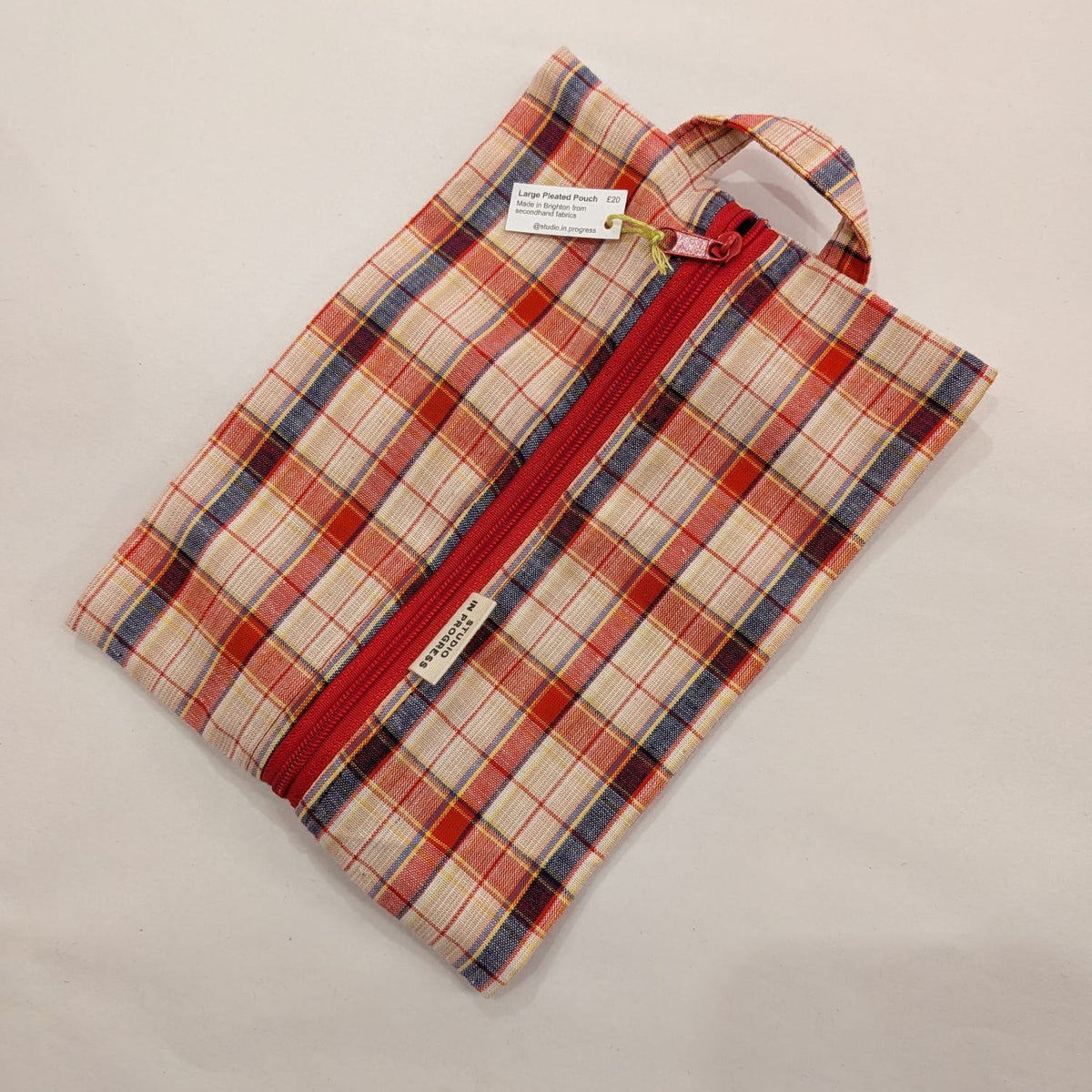 A small zippered pouch with a red and cream plaid pattern, featuring a red zipper, a fabric loop handle, and two small white tags, shown on a light beige background.