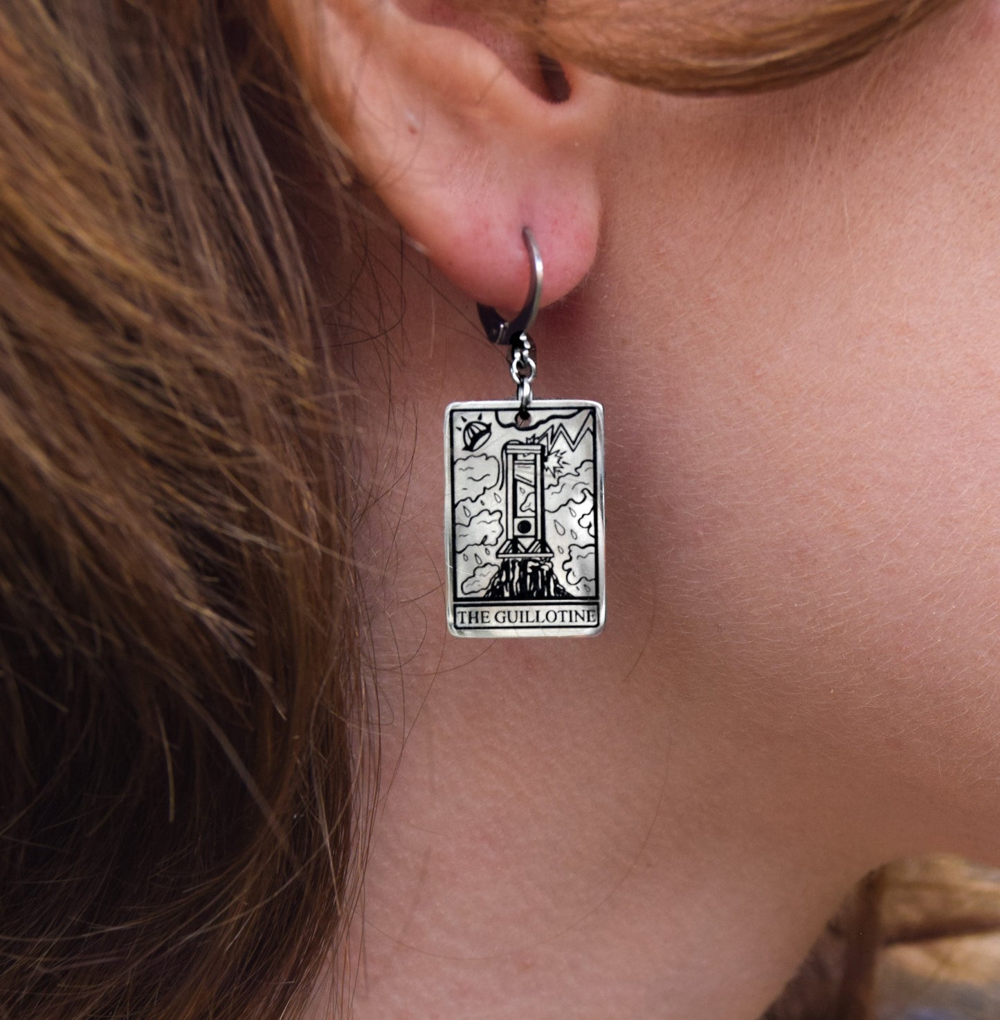 Silver earring with 'The Guillotine' tarot card design worn by a person.