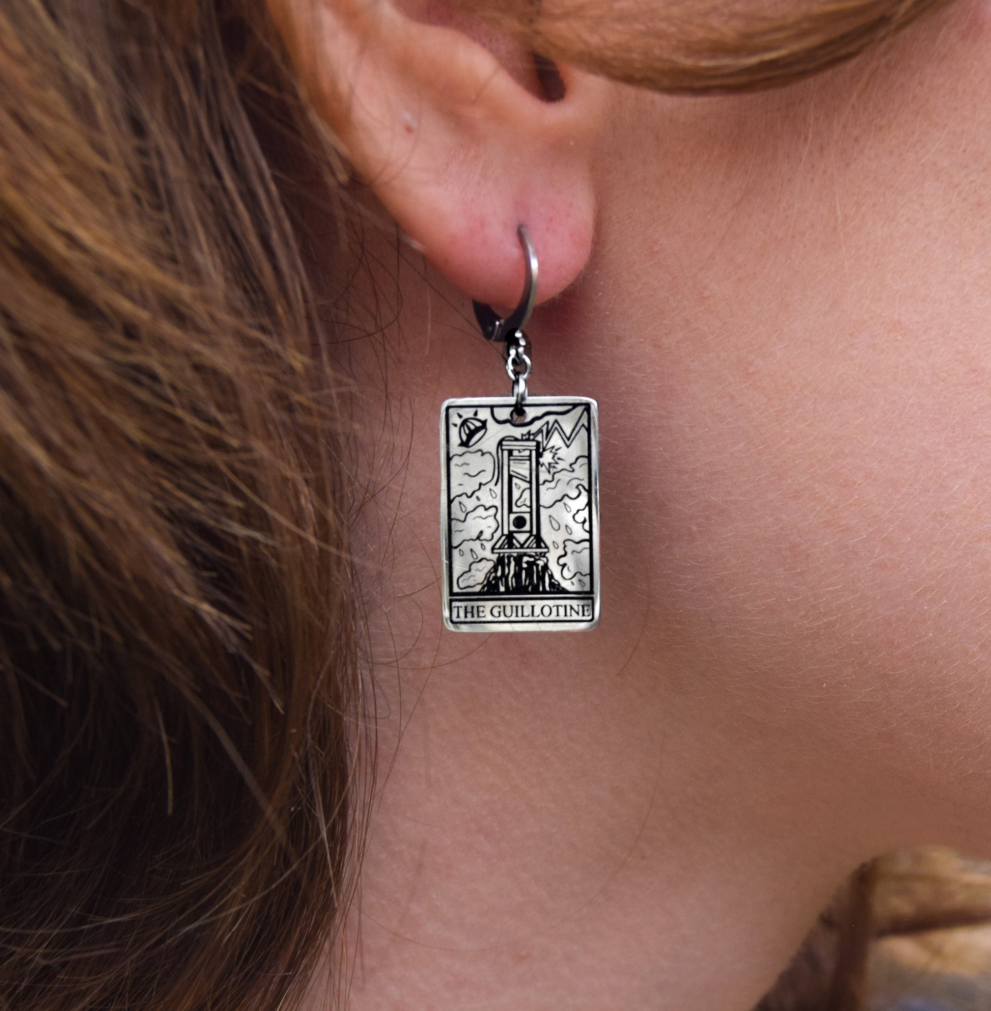 Silver earring with 'The Guillotine' tarot card design worn by a person.