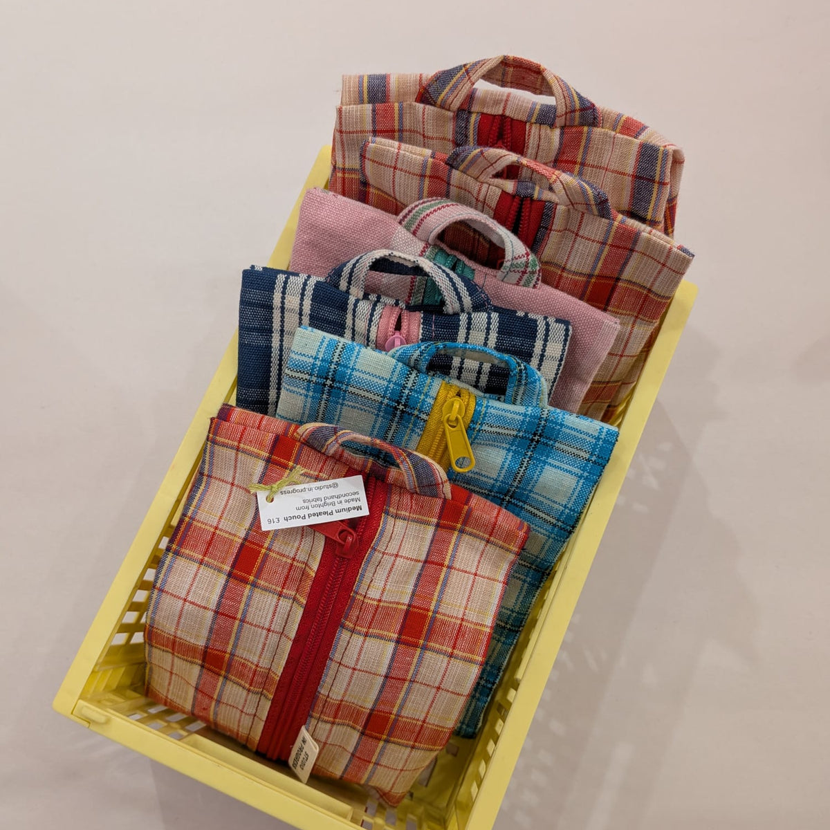 A yellow plastic storage basket filled with multiple small fabric zippered pouches or bags in plaid patterns, photographed against a plain light background.