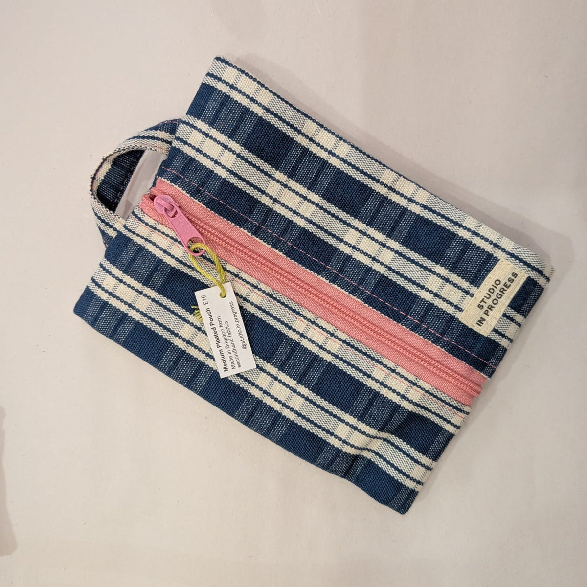 A small zippered pouch with a blue, white, and pink plaid pattern, featuring a pink zipper, a fabric handle, and a hanging product tag, displayed on a light beige surface.