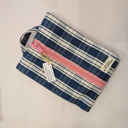 A small zippered pouch with a blue, white, and pink plaid pattern, featuring a pink zipper, a fabric handle, and a hanging product tag, displayed on a light beige surface.