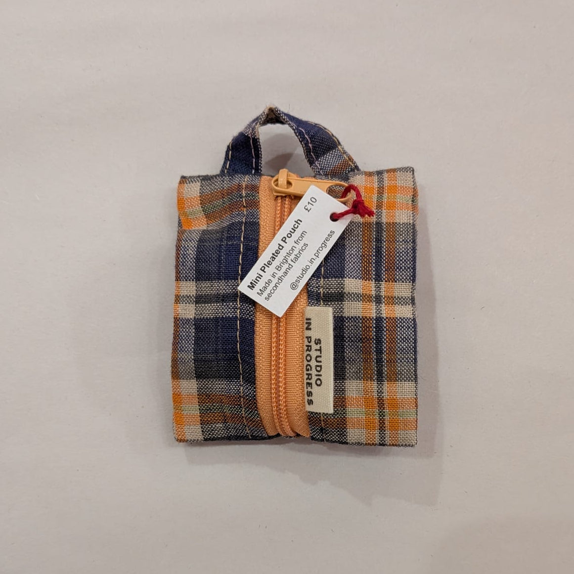 A small zippered fabric pouch with a plaid pattern in navy blue, orange, and beige, featuring a fabric loop handle and a white paper tag attached. The tag reads 'STUDIO by PIERRE' and 'MADE IN FRANCE'.