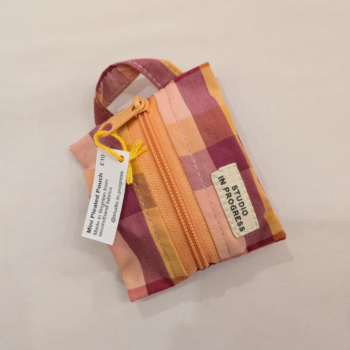 A small zippered fabric pouch with a multicolor plaid pattern in pink, orange, yellow, and purple, featuring a white brand label that reads 'MY STITCHES STUDIO' and a product tag.