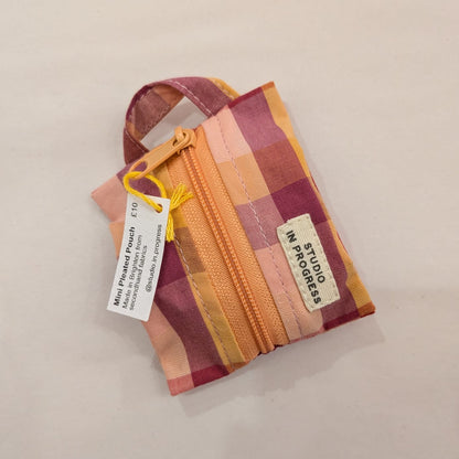 A small zippered fabric pouch with a multicolor plaid pattern in pink, orange, yellow, and purple, featuring a white brand label that reads 'MY STITCHES STUDIO' and a product tag.