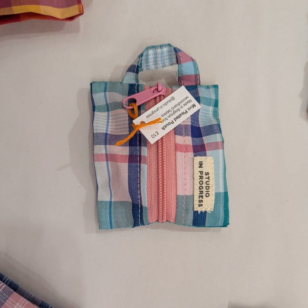 A small zippered fabric pouch with a plaid pattern in blue, pink, and white, featuring a pink zipper pull and a hanging paper tag that reads 'STUDIO IN PROGRESS'.