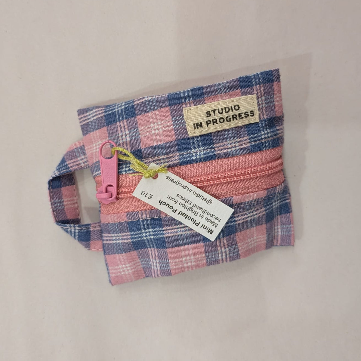 A small zippered pouch in a pink and blue plaid fabric, featuring a pink zipper pull, a yellow-green drawstring, and a white paper tag that reads 'Mini Plaid Pouch' and 'STUDIO IN PROGRESS'.