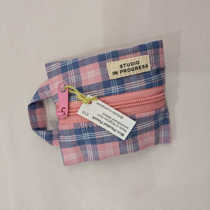 A small zippered pouch in a pink and blue plaid fabric, featuring a pink zipper pull, a yellow-green drawstring, and a white paper tag that reads 'Mini Plaid Pouch' and 'STUDIO IN PROGRESS'.