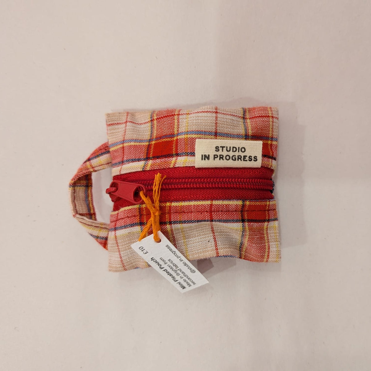 A small fabric zippered pouch with a red zipper and a cream label reading 'STUDIO IN PROGRESS', photographed on a plain off-white background.