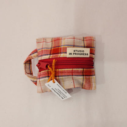 A small fabric zippered pouch with a red zipper and a cream label reading 'STUDIO IN PROGRESS', photographed on a plain off-white background.