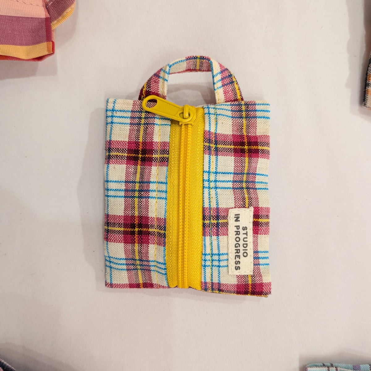A small fabric zippered pouch with a multicolor plaid pattern, featuring a bright yellow zipper and a white fabric label that reads 'STOCKMAN IN SUGAR DADDY'.