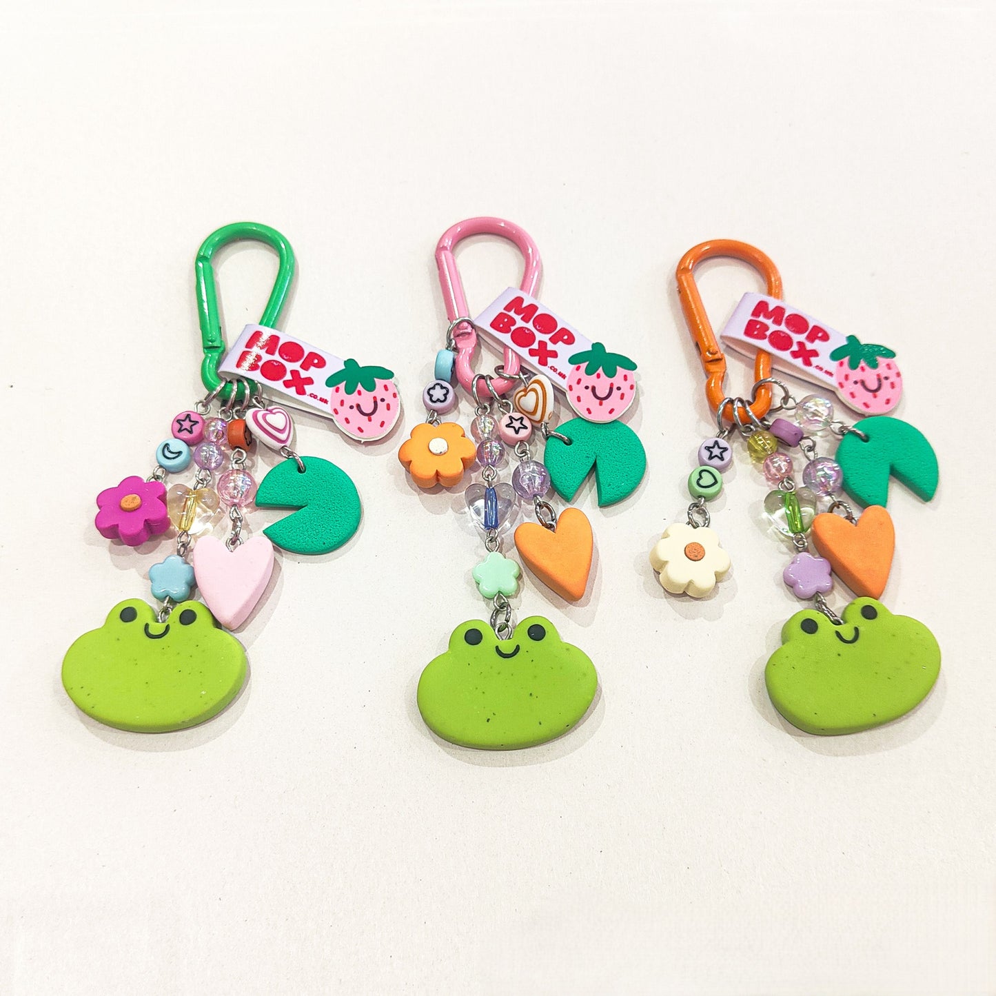 A keychain featuring charms shaped like frog faces, flowers, and hearts. Three are laid side by side in green, pink and orange colour ways.