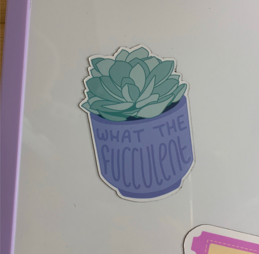 A fridge magnet with a light green succulent plant in a lavender-blue pot with the text 'WHAT THE FUCCULENT' written on it, shown on a white surface.
