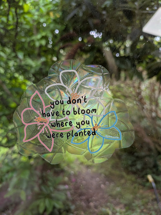 A translucent flower-shaped sun catcher with the quote 'you don't have to bloom where you were planted' in black script, decorated with pink, white, and blue flowers.