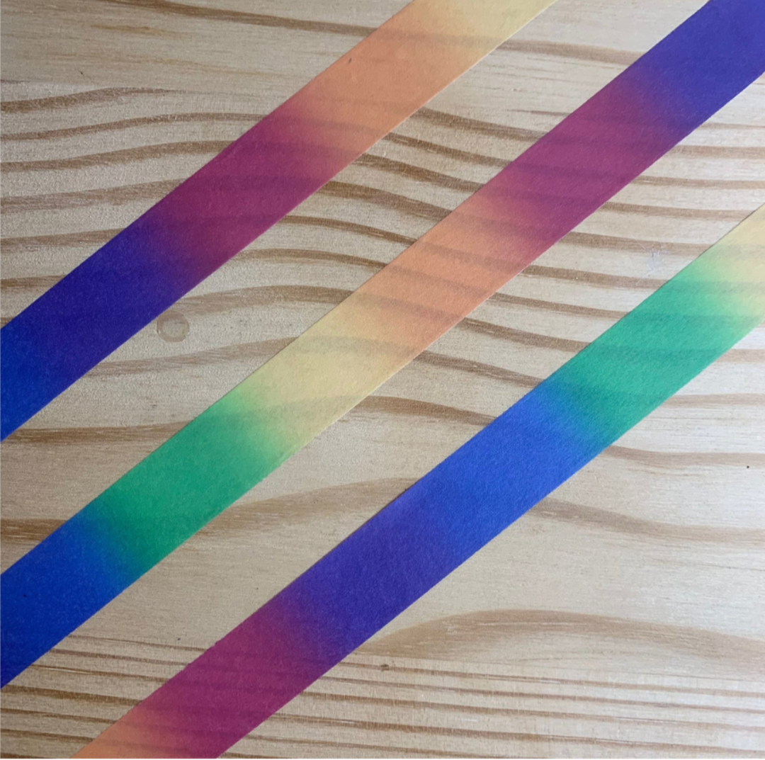 Three diagonal strips of washi tape with a rainbow gradient pattern, placed on a light wood grain surface.